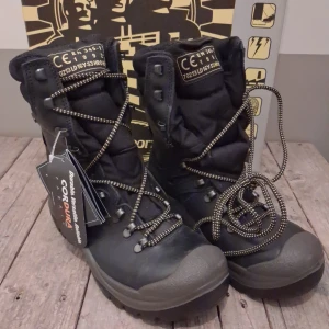 Svarta Grisport kängor med snörning - Svarta kängor från Grisport med robust snörning och grov platt sula. Tillverkade i slitstark Cordura och skinn, med Sympatex support system för extra komfort. Märkta med säkerhetsstandard och har detaljer i gult och orange på sula och etiketter. Ny pris 2300kr