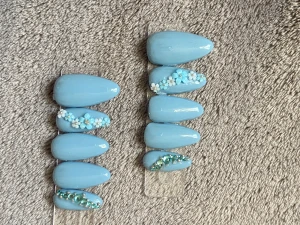 Blåa lösnaglar med blommor och stenar - Snygga mandelformade lösnaglar i ljusblått med 3D-blommor och glittrande stenar. Några naglar har små vita och blå blommor, andra är dekorerade med blå och silverfärgade stenar. Perfekt för dig som vill ha en unik och kreativ nageldesign.