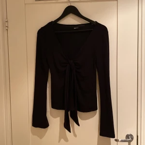 Svart långärmad topp med knyt Gina Tricot - Svart långärmad topp från Gina Tricot med djup v-ringning och snygg knytning framtill. Modellen är figurnära och har långa ärmar med lätt utsvängd form. Perfekt för att lyfta en enkel outfit med en trendig detalj.
