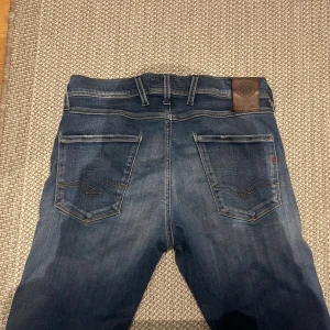 Replay Anbass jeans blå, storlek 29 - Snygga Replay Anbass jeans i klassisk blå tvätt med diskreta slitningar och kontrastsömmar. Modellen har fem fickor och normal passform. Märkespatch baktill och detaljerad brodering på bakfickorna. Perfekt för dig som gillar stilrena jeans med lite edge.