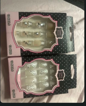 Sara Nail lösnaglar med dekoration - Två förpackningar med  lösnaglar vardera från Sara Nail. Ena setet har transparenta naglar med silverglittriga stenar, andra har vita blommönster på transparent bas. Naglarna är långa och har elegant form, perfekta för att skapa en snygg manikyr hemma.