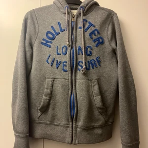 Grå hoodie från Hollister med blå text - Grå hoodie från Hollister med blå broderad text på framsidan. Tröjan har huva med snörning, två fickor framtill och en blå dragkedja. Tillverkad i mjuk bomullsmix, perfekt för chill dagar. Långärmad och med ribbade muddar för extra komfort.