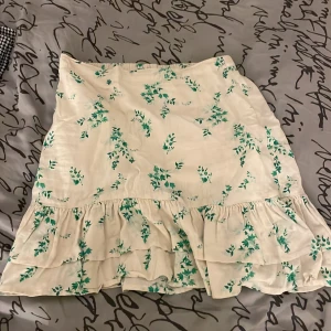 Vit blommig volangkjol från Lindex - Supersöt vit kortkjol från Lindex med grönt blommigt mönster och volanger nedtill. Kjolen har elastisk midja och är gjord i mjukt, lätt material som känns skönt mot huden. Perfekt för dig som gillar en fräsch och somrig stil. 🤗🤗