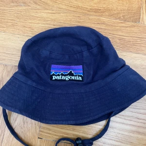 Mörkblå bucket hat från Patagonia - Mörkblå bucket hat från Patagonia med klassisk broderad logga framtill i lila, blått och rött. Hatten har rund form och dragsko under hakan. 