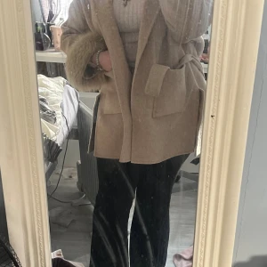 Beige kappa med pälsdetaljer - Säljer en beige kappa med fluffiga pälsdetaljer vid ärmsluten och stora fickor framtill. Jackan har en omlottstil med bälte i midjan och är perfekt för dig som vill ha en chill och snygg look under höst och vinter. Den passar upp till L/XL men passar i princip alla.