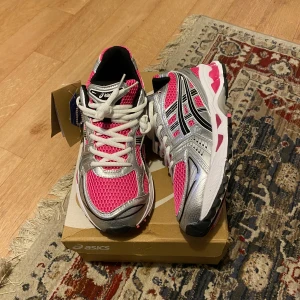 Asics rosa och silver sneakers strl 42 - Snygga Asics sneakers i storlek 42 med mesh-överdel i stark rosa och silverdetaljer. Skorna har vita snören, svart innersida och coola svarta och rosa detaljer på sulan. Perfekta för dig som gillar sportig stil och vill sticka ut med färg.