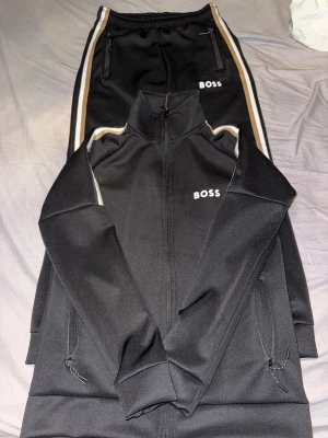 Svarta BOSS mjukisbyxor med revärer - Svarta mjukisbyxor från BOSS med vita och beige revärer längs sidorna. Byxorna har resår i midjan, dragkedjefickor och BOSS-logga på låret. Snygg sportig look, perfekta för chill eller träning.priset kan diskutera