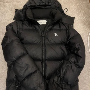 Svart pufferjacka från Calvin Klein Jeans - Svart pufferjacka från Calvin Klein Jeans med stor huva och dragkedja framtill. Jackan har broderad CK-logga på bröstet och en stor patch på ärmen. Yttermaterialet är 100% återvunnen nylon och fodret är 100% återvunnen polyester. Vattenavvisande och fylld med ansvarsfullt dun. Skick 8,5/10. Skriv om du har fler frågor. Storlek XS