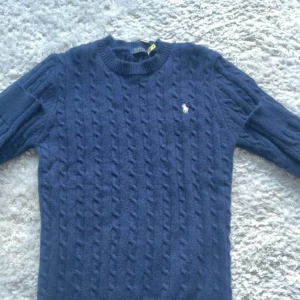 Marinblå stickad tröja Polo Ralph Lauren - Snygg marinblå stickad tröja från Polo Ralph Lauren med klassiskt kabelmönster och broderad vit logga på bröstet. Rund hals och långa ärmar, perfekt för dig som gillar stilren och tidlös design. Tröjan är i mjuk ull och har ribbade muddar vid hals, ärmslut och nederkant.