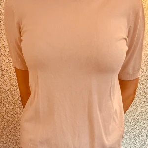 Ljusrosa stickad topp med kort ärm - Snygg ljusrosa stickad topp med korta ärmar och ribbad halsringning. Baksidan har en dekorativ söm och en liten metallring vid nacken som detalj. 