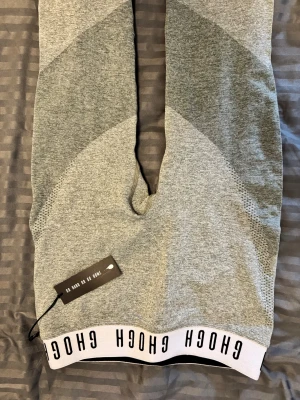 Grå träningsleggings från GHOGH M - Snygga grå leggings från GHOGH med bred vit resår i midjan med svart text 'GO GH OR GO HOME'. Materialet är en mix av nylon, polyester och elastan, vilket gör dem stretchiga och bekväma. Perfekta för gymmet eller sport. Diskret logga på benet.