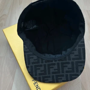 Svart Fendi keps med FF-mönster - Snygg svart keps från Fendi med det klassiska FF-logomönstret över hela kepsen. Kepsen har böjd skärm och justerbar öppning bak med Fendi Roma-broderi. Materialet är slitstark bomull och polyester, perfekt för dig som gillar exklusiva accessoarer.