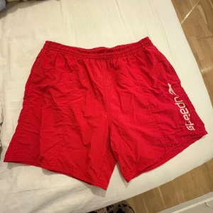 Röda badshorts från Speedo - Säljer ett par röda badshorts från Speedo med vit logga på sidan. De har elastisk midja med snörning och meshfoder på insidan. Shortsen har en bakficka med knapp och är tillverkade i lätt syntetmaterial, perfekta för stranden eller poolen.
