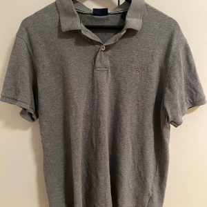 Grå pikétröja från GANT - Snygg grå pikétröja från GANT med klassisk krage och korta ärmar. Tröjan har två knappar vid halsen och en liten broderad logga på bröstet. Tillverkad i mjuk bomull som är skön mot huden. Perfekt för dig som gillar stilren och enkel design.