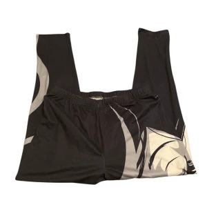 Svarta leggings med ansiktsmotiv - Unika svarta leggings med ett stort grafiskt ansiktsmotiv i grått och vitt på framsidan, samt röda läppar som sticker ut. Byxorna har elastisk midja och är tillverkade i mjukt stretchmaterial som sitter skönt. 