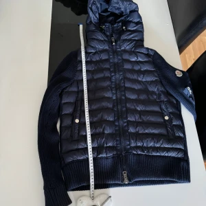 Moncler cardigan  - Säljer en mörkblå pufferjacka från Moncler med stickade ärmar och ribbade muddar. Jackan har huva, tvåvägsdragkedja, två fickor med tryckknappar och Moncler-logga på ärmen. Perfekt för dig som vill ha en snygg och sportig look.