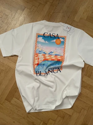 Vit Casa Blanca t-shirt med tryck - Snygg vit t-shirt från Casa Blanca i 100% ekologisk bomull. Framtill finns ett färgstarkt tryck med sol, moln och hav i orange och blå toner samt logotypen. Klassisk rund hals och korta ärmar. Perfekt för dig som gillar stilrena plagg med unik detalj.