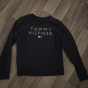 Mörkblå Tommy Hilfiger sweatshirt - Snygg mörkblå sweatshirt från Tommy Hilfiger med stort tryck på bröstet och klassisk logga i vitt, rött och blått. Tröjan har rund hals, långa ärmar och ribbade muddar. Perfekt för dig som gillar stilrena märkesplagg och vill ha en enkel, clean look.