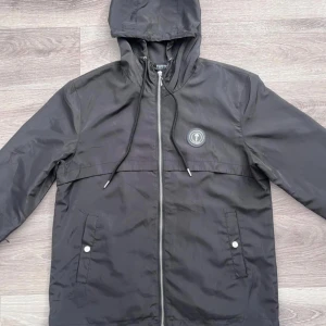 Svart vindjacka från Trapstar - Svart vindjacka från Trapstar med huva och dragkedja framtill. Jackan har logotyp på bröstet och broderad Trapstar-text på ryggen. Två fickor med knappstängning och justerbar snörning vid huvan. Materialet är lätt och syntetiskt, perfekt för streetwear.