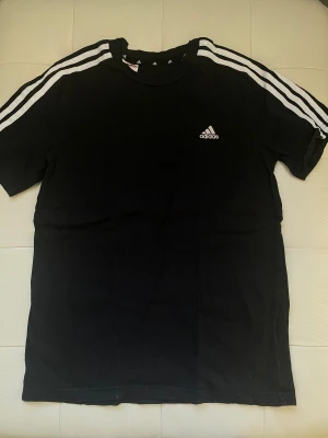 Svart Adidas t-shirt med vita ränder - Svart t-shirt från Adidas med klassiska vita ränder längs axlarna och ärmarna. Adidas-logga på bröstet. Skön passform och tillverkad i mjuk bomull. Perfekt för en sportig och avslappnad stil.