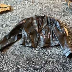 Svart croppad vintage skinnjacka - Säljer en svart croppad skinnjacka  med coola detaljer och breda slag, stora puffiga axlar. Jackan har långärmade ärmar och knappar framtill. Insidan har ett mönstrat foder med grafiska former i brons och rosa toner. Perfekt för dig som vill ha en edgy look.