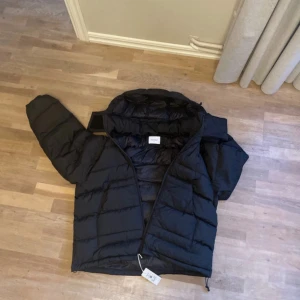 Svart pufferjacka från vailent - Svart pufferjacka från vailent med huva och dragkedja framtill. Jackan har breda, quiltade sektioner och en loose passform som ger en chill vibe. Perfekt för kalla dagar och har justerbar dragsko nedtill. Enkel, stilren och riktigt skön. Storlek M skick 10/10 nypris 1499