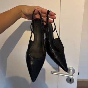Svarta pumps klackar med spetsig tå och klack - Säljer ett par svarta pumps / klackar med glansig finish och spetsig tå. Skorna har en smal rem runt hälen med spänne och en elegant låg klack. Köpta på asos men använda inte längre!