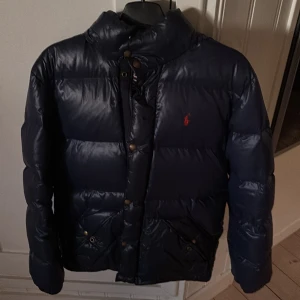 Mörkblå dunjacka från Polo Ralph Lauren - Snygg mörkblå dunjacka från Polo Ralph Lauren med röd broderad logga på bröstet. Jackan har hög krage, tryckknappar och dragkedja framtill. Perfekt för kalla dagar och har en klassisk, puffad look som är både trendig och bekväm.
