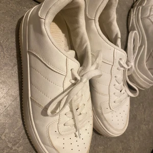 Vita klassiska sneakers med snörning - Säljer ett par helvita sneakers med stilren design och snörning. Skorna har rund tå, perforerade detaljer framtill och är gjorda i syntetmaterial med textilfoder. Perfekta för dig som gillar en clean och enkel look till vardags.