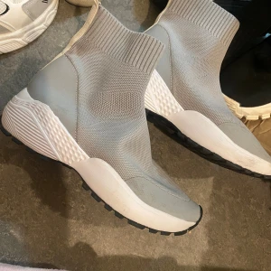 Grå stickade sock-sneakers - Säljer ett par grå stickade sock-sneakers med högt skaft och vit chunky sula. Skorna har en modern, strumpliknande passform och svart yttersula för extra grepp. Perfekta för dig som gillar en sportig och trendig look.
