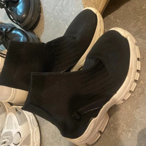 Svarta sock-sneakers med vit sula - Säljer ett par svarta sock-sneakers med chunky vit sula. Skorna är stickade i syntetmaterial och har en hög, ribbad skaft som sitter tight runt foten. Perfekta för dig som gillar streetstyle och vill ha något bekvämt men ändå trendigt.