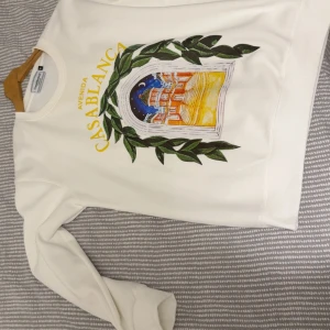 Vit sweatshirt från Casablanca - Vit sweatshirt från Casablanca med färgglatt tryck på framsidan som föreställer en byggnad och gröna blad. Tröjan har rund hals, långa ärmar och ribbade muddar. Perfekt för dig som gillar snygga prints och vill sticka ut lite extra.
