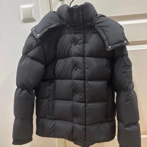 Svart Moncler pufferjacka med huva - Svart pufferjacka från Moncler med avtagbar huva och klassisk logga på ärmen. Jackan har quiltad design, knappar framtill och praktiska fickor. Perfekt för kalla dagar och har en riktigt snygg, puffig look som sticker ut.