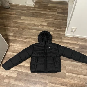 Svart Nike pufferjacka Therma-Fit XS - Svart pufferjacka från Nike i modellen Therma-Fit. Jackan har huva, dragkedja framtill och två sidofickor. Den är quiltad och har en clean look med diskret Nike-logga på bröstet. Perfekt för kalla dagar och har en skön, loose passform.