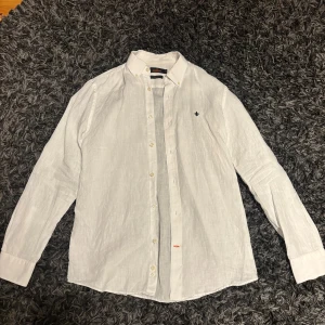 Vit linneskjorta från Morris - Vit skjorta från Morris i mjukt linne med klassisk krage och knappar framtill. Långärmad modell med diskret blå broderad logga på bröstet. Perfekt för dig som gillar stilrena och fräscha plagg med snygga detaljer.