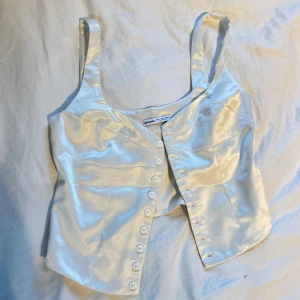 Vit satin korsett från Bershka - Säljer en vit korsett-topp från Bershka i glansigt satinmaterial. Toppen har breda axelband, figurnära passform och knappar framtill🌸