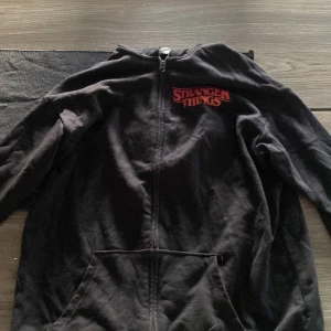 Svart Stranger Things hoodie från H&M - Svart hoodie från H&M med Stranger Things-tryck i rött på bröstet och en stor färgglad bild av Eleven på ryggen. Tröjan har huva, dragkedja framtill och praktisk ficka. Perfekt för dig som älskar serien och vill ha en cool streetstyle.