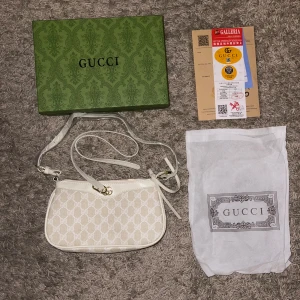 Vit axelväska från Gucci med GG-mönster - Snygg axelväska från Gucci i vitt och beige med klassiskt GG-mönster. Väskan har guldfärgade detaljer, dragkedja och justerbar axelrem. Tillverkad i canvas och skinn med innerfack i ljust material. Kommer med originalbox och dustbag.