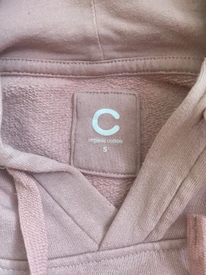 Ljusrosa hoodie i ekologisk bomull - Säljer en ljusrosa hoodie från Cubus i ekologisk bomull. Tröjan har huva med snörning, stor magficka och långa ärmar med ribbade muddar. 