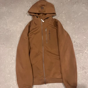 Brun hoodie från Sail Racing med dragkedja - Snygg brun hoodie från Sail Racing med huva och hel dragkedja framtill. Tröjan har en bröstficka med logga och långa ärmar. Perfekt för dig som gillar stilren design och vill ha något bekvämt och coolt i garderoben.