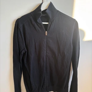 Svart cardigan jacka från Selected Homme - Svart cardigan jacka från Selected Homme med hel dragkedja framtill och hög krage. Jackan är stickad och har långärmade ärmar, perfekt för dig som gillar stilren och enkel design. Passar dig som vill ha en snygg och bekväm jacka till vardags.