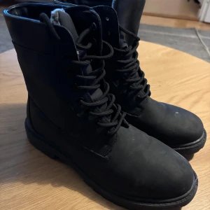 Svarta kängor med snörning/ stövlar vinter - Säljer ett par svarta kängor med grov sula och snörning framtill. De har rund tå och är fodrade med mjukt material på insidan för extra värme. Utsidan är i matt skinn och mocka, vilket ger en cool och robust look. Perfekta för höst och vinter.