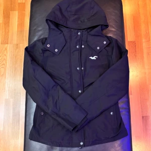 Svart vindjacka från Hollister M - Svart vindjacka från Hollister med huva och tryckknappar framtill. Jackan har en stilren design med Hollister-logga på bröstet och praktiska fickor med knappstängning. Perfekt för blåsiga dagar och enkel att matcha med din vardagsstil.