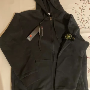 Svart hoodie från Stone Island - Svart hoodie från Stone Island med klassisk logotyp-patch på ärmen, dragkedja framtill och snörning vid huvan. Tröjan har ribbade muddar och är tillverkad i mjuk bomull. Perfekt för dig som gillar streetwear och vill ha en clean look.