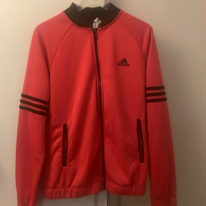 Röd Adidas bomberjacka M - Snygg röd bomberjacka från Adidas med svarta detaljer och klassiska tre ränder på ärmarna. Jackan har dragkedja framtill, två fickor på utsidan och även 2 inne ficor och svart krage. Den är som helt ny inget är trasigt 