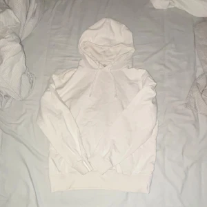 Vit hoodie med huva och snörning - Säljer en stilren vit hoodie med huva och snörning. Tröjan har långärmade ärmar och ribbade muddar vid ärmslut och nederkant. Perfekt för chill dagar och enkel att matcha med jeans eller joggers.