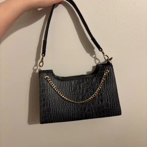 Svart axelväska med kedjedetalj - Stilren svart axelväska i croco-mönstrad syntet med guldfärgad kedja framtill. Väskan har avtagbart axelband och stängs med dragkedja. Perfekt storlek för det viktigaste och ger en cool vibe till din outfit.