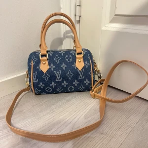  denim handväska - Snygg handväska i blå denim med klassiskt monogrammönster. Väskan har ljusbruna skinnhandtag och avtagbar axelrem i samma färg. Guldiga metalldetaljer och dragkedja upptill. Perfekt storlek för det viktigaste och en riktigt trendig accessoar.