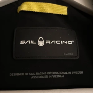 Svart vindjacka Sail Racing L - Svart vindjacka från Sail Racing i storlek Large. Jackan har huva, hel dragkedja framtill och snygga logoprints på bröstet, ärmarna och ryggen. Materialet är slitstark syntet som står emot vind och väta, perfekt för aktiva dagar utomhus.