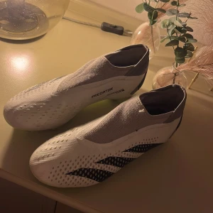 Adidas Predator vita fotbollsskor - Adidas Predator fotbollsskor i vitt med svarta detaljer och limegröna dobbar. Skorna har en strumpliknande form utan snörning, vilket ger en tight och modern look. Ovandelen är tillverkad i syntetmaterial med ventilerande mesh och mönstrad struktur för extra grepp på bollen.